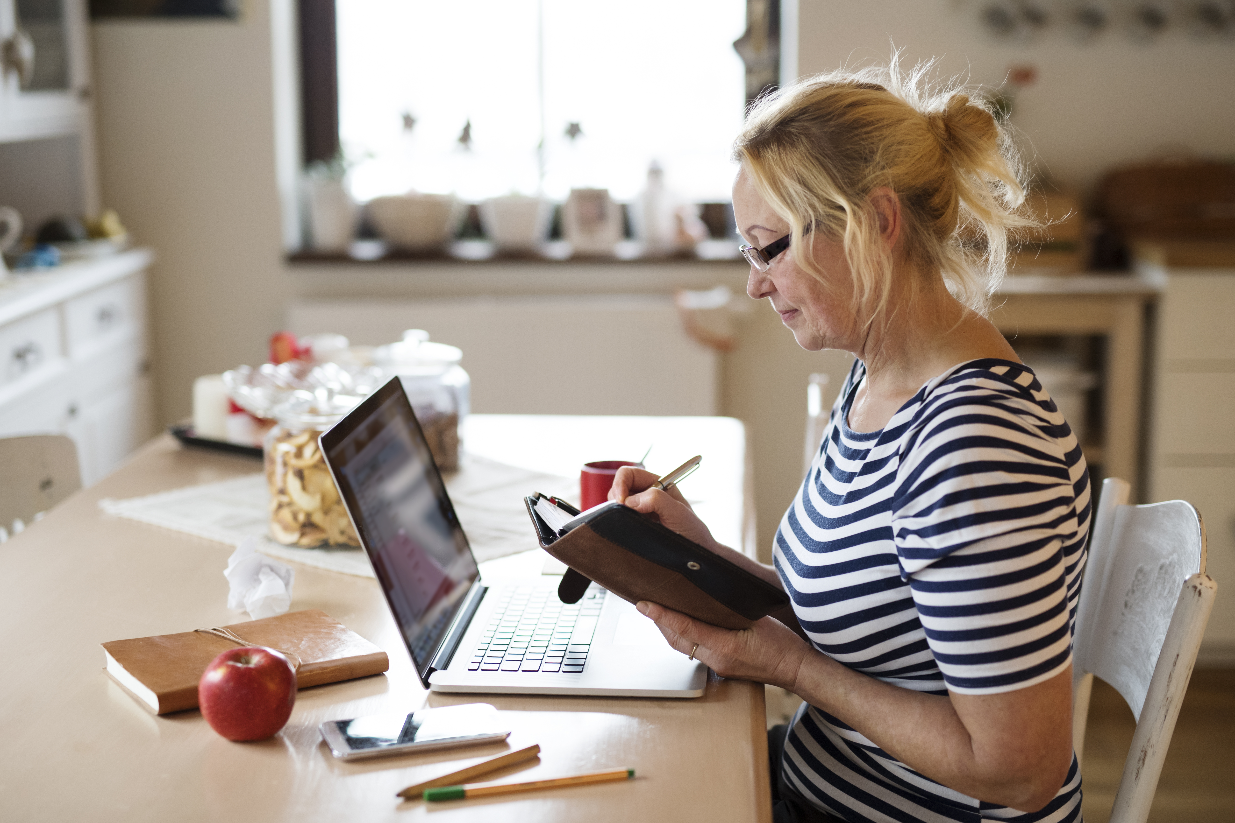 Frau mit Brille arbeitet am Laptop und notiert Termine in einen Kalender am Küchentisch.