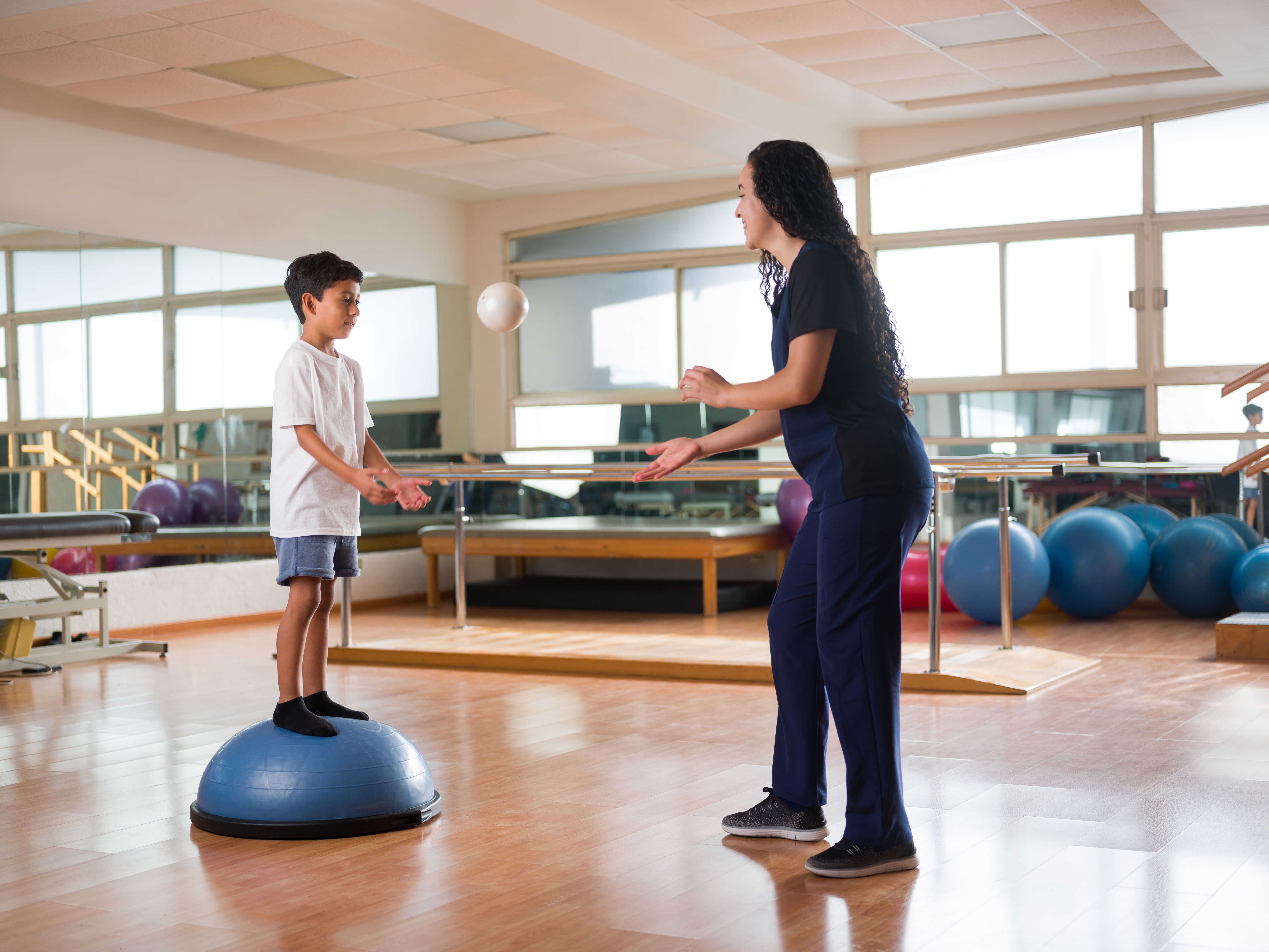 Junge balanciert auf Bosu-Ball und spielt Ball mit Physiotherapeutin in Therapieraum